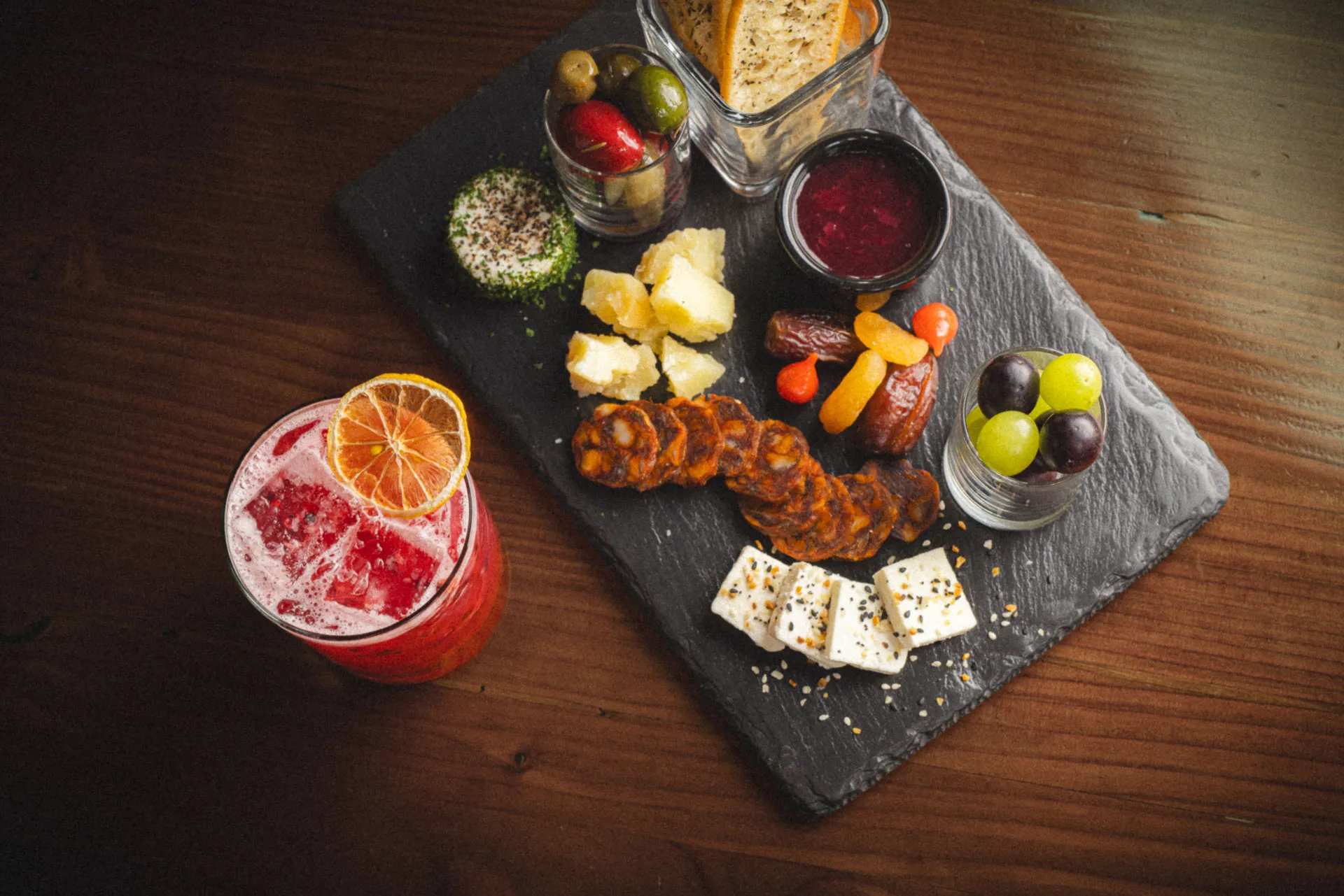 Overhead view of a gourmet charcuterie board featuring Everything Feta, Local Goat Cheese, Spanish Manchego, Cabernet Jam, Chef's Selection of Olives, Iberico Chorizo, and Crostini, complemented by a blackberry lemonade to the left.